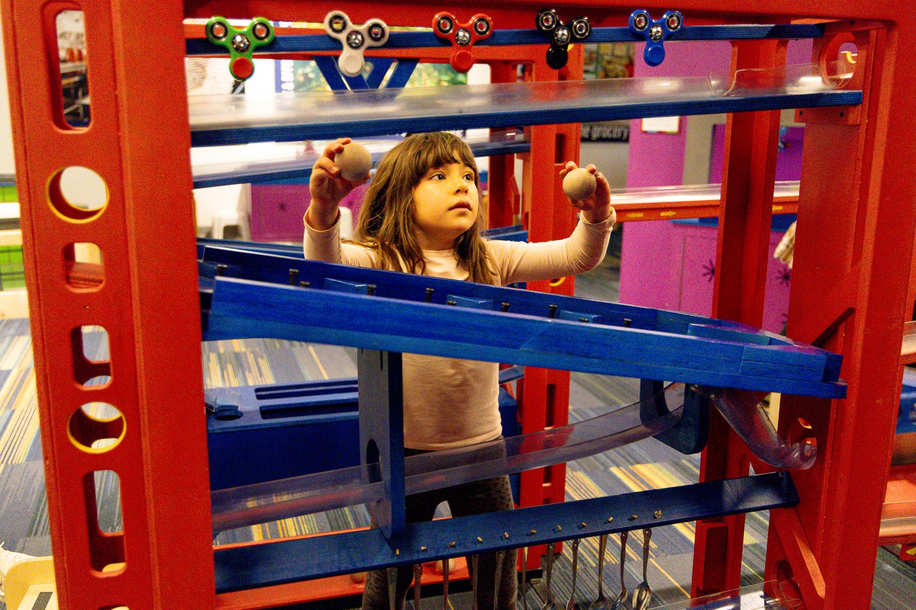 a young girl stands among several sets of towers and ramps, putting balls into the ramps