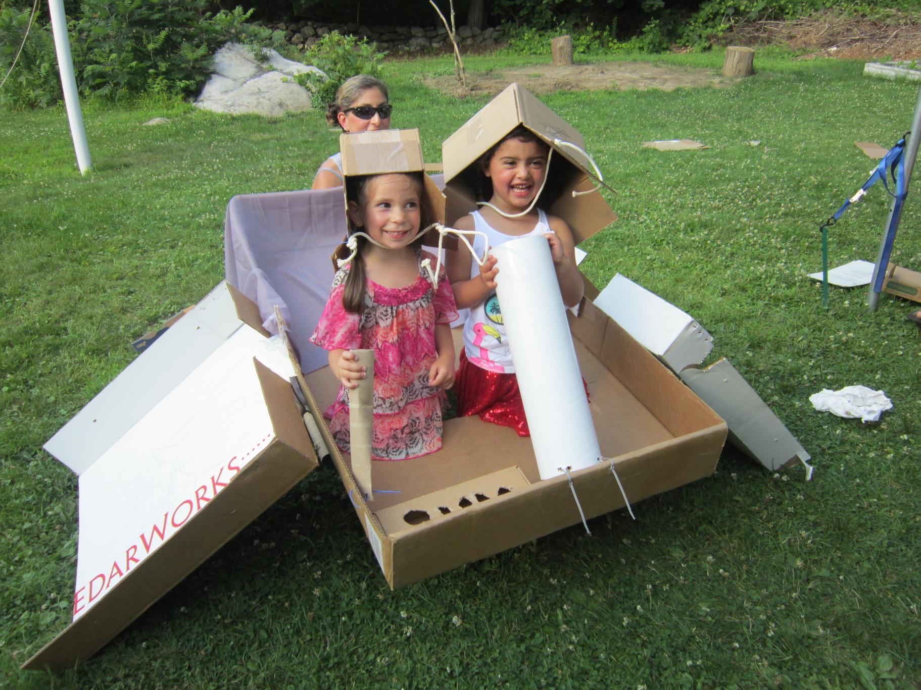two young girls sit in a cardboard "plane" that they created out of boxes