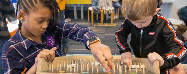 two children, an older girl and younger boy, play together at a tabletop pegboard