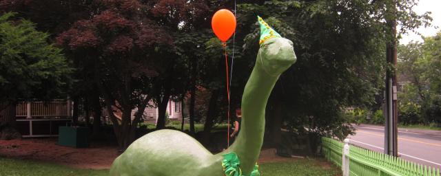 a large dinosaur statue has balloons tied to it and wears a party hat