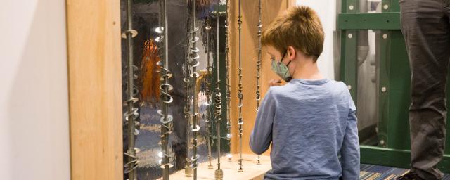 a boy kneels on a floor in front of a mirrored wall with metal washers running down vertical rods in front of the mirror