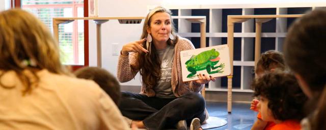 a woman sits on the floor reading a picture book to children, using sign language